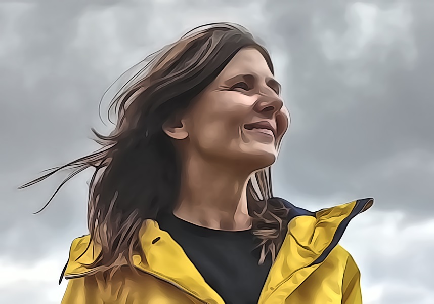 Emma ve žluté bundě, usmívá se, vítr jí vlaje ve vlasech. Za ní jsou vidět mraky
